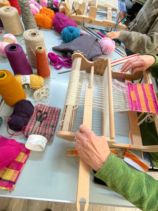 Person weaving with a loom surrounded by colorful yarn and fabric samples.